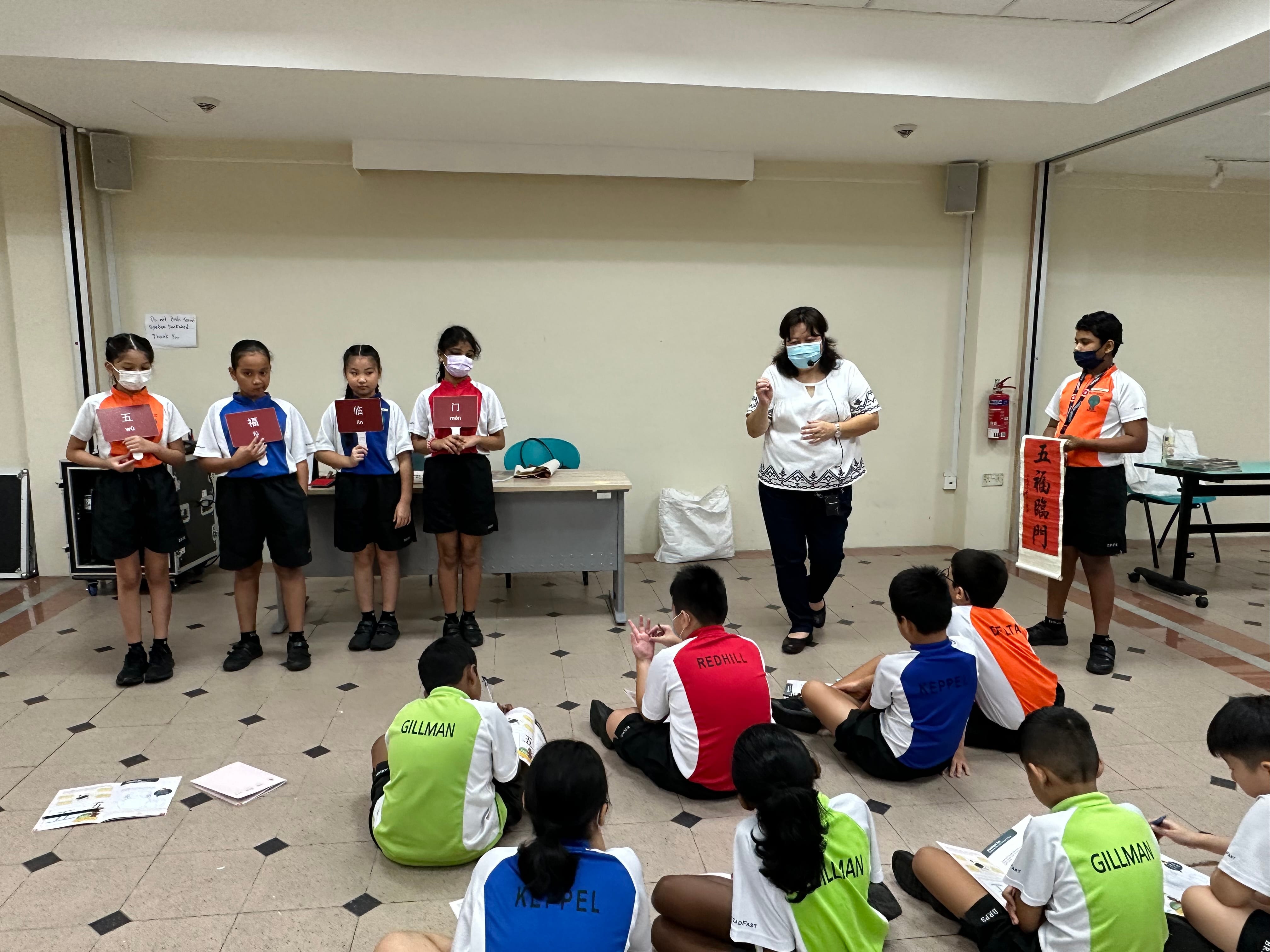 A teacher and students stand holding Chinese character cards while other children sit on the floor watching in a classroom.